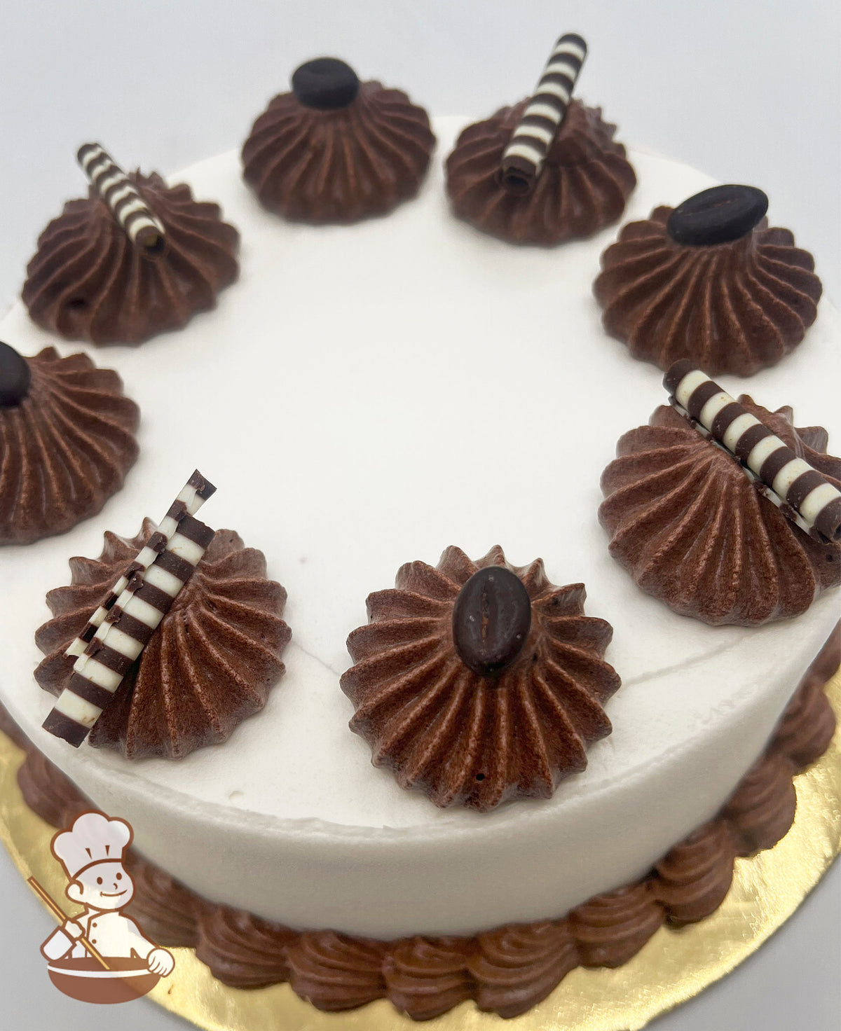 Round cake with smooth white frosting, topped with brown buttercream swirls, striped chocolate sticks, and coffee bean decorations.