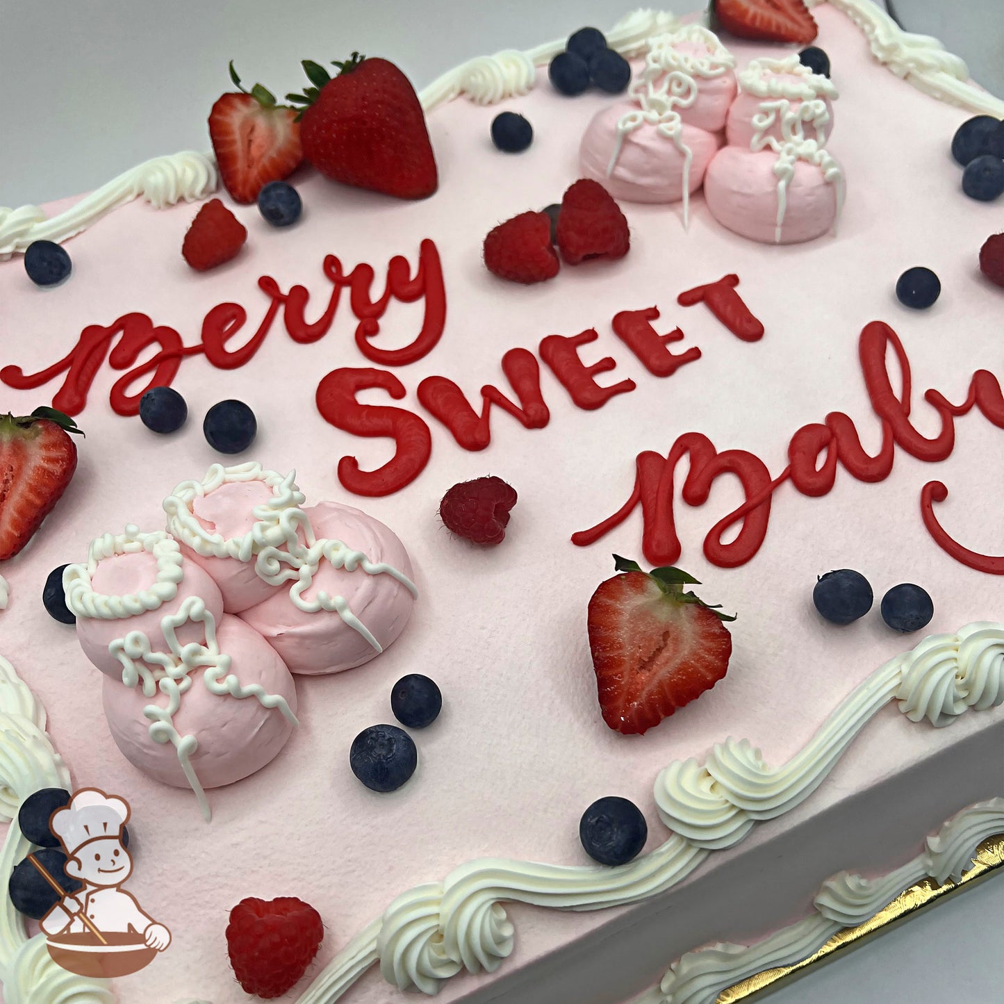 Pink sheet cake with white buttercream borders, decorated with fresh strawberries, blueberries, raspberries, and pink buttercream baby booties with white piping details.