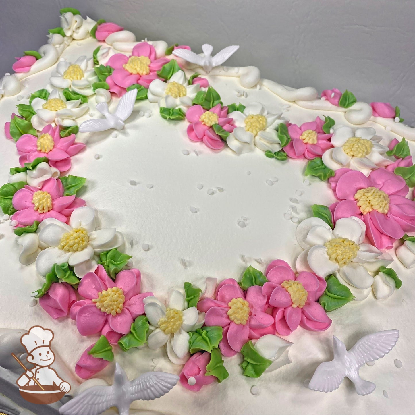 Heart-shaped buttercream floral design with pink and white blossoms, green leaves, and plastic white dove decorations on a sheet cake.