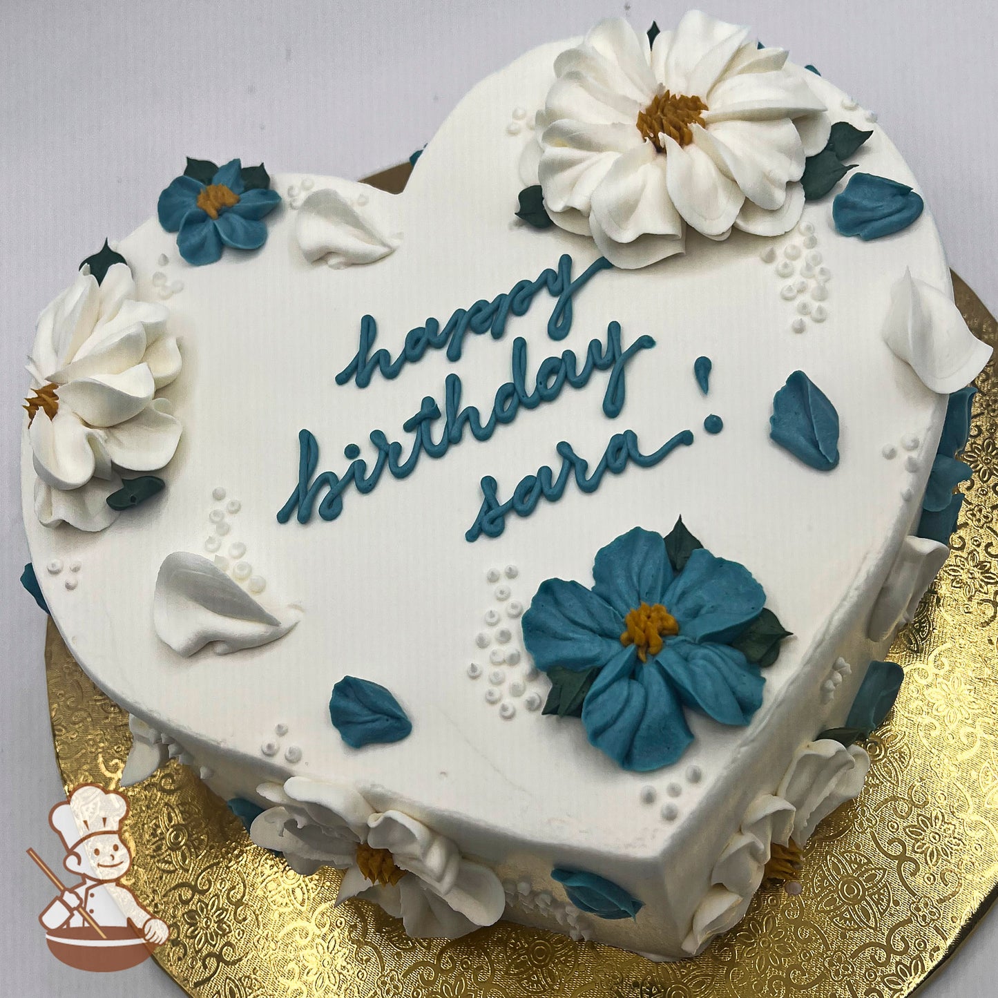 Heart-shaped cake with white icing, decorated with white and blue blossoms, and petals.