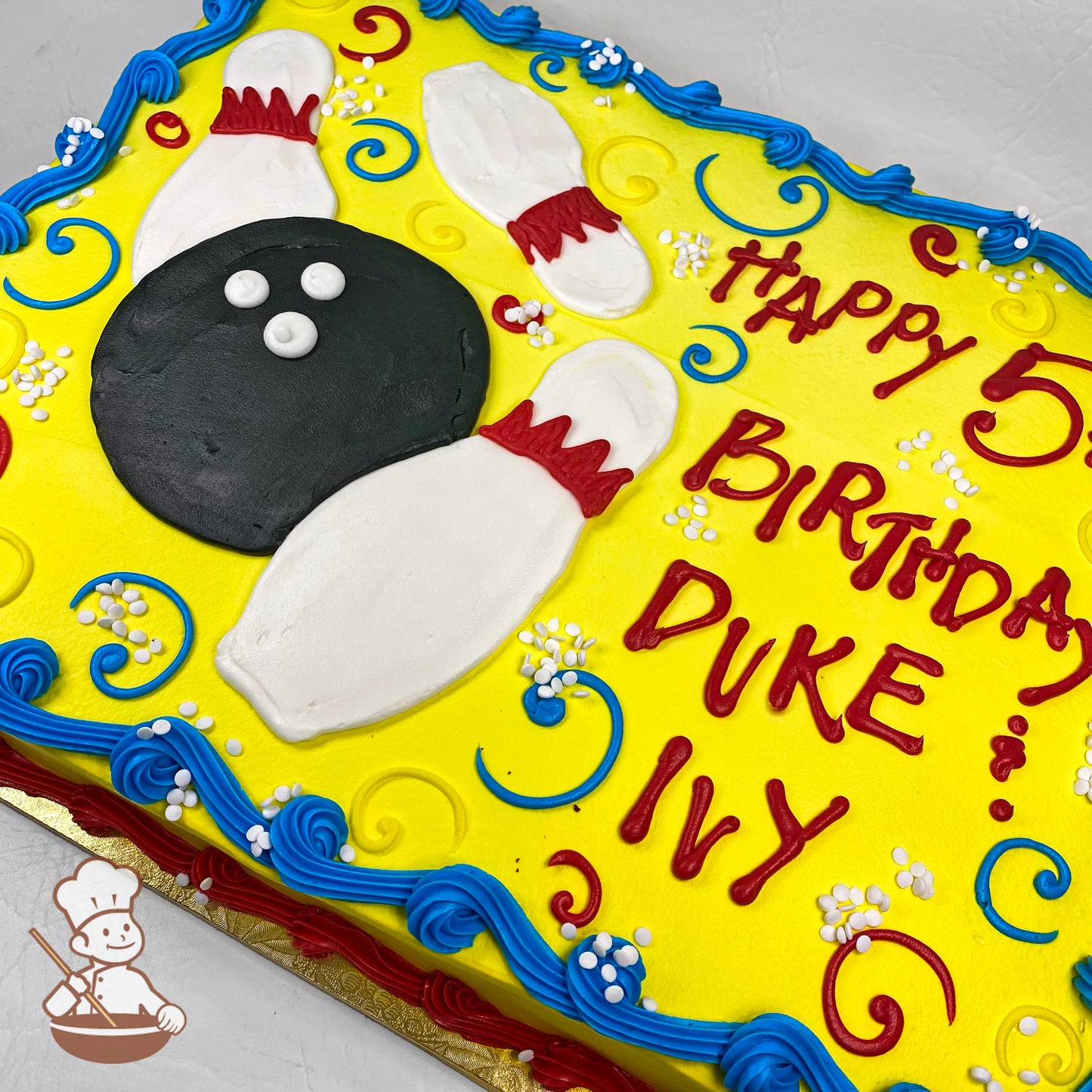 Sheet cake with yellow buttercream, decorated with a black bowling ball, three white bowling pins with red accents, blue and red swirls, and small white flowers.