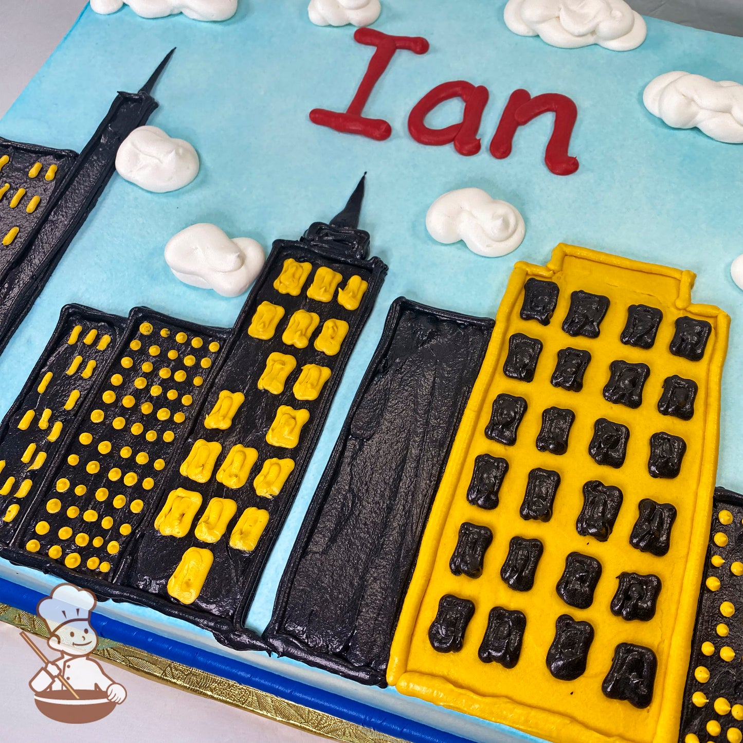 Sheet cake with buttercream decorations of a city skyline at day, featuring tall black and yellow buildings, fluffy white clouds, and a light blue sky.