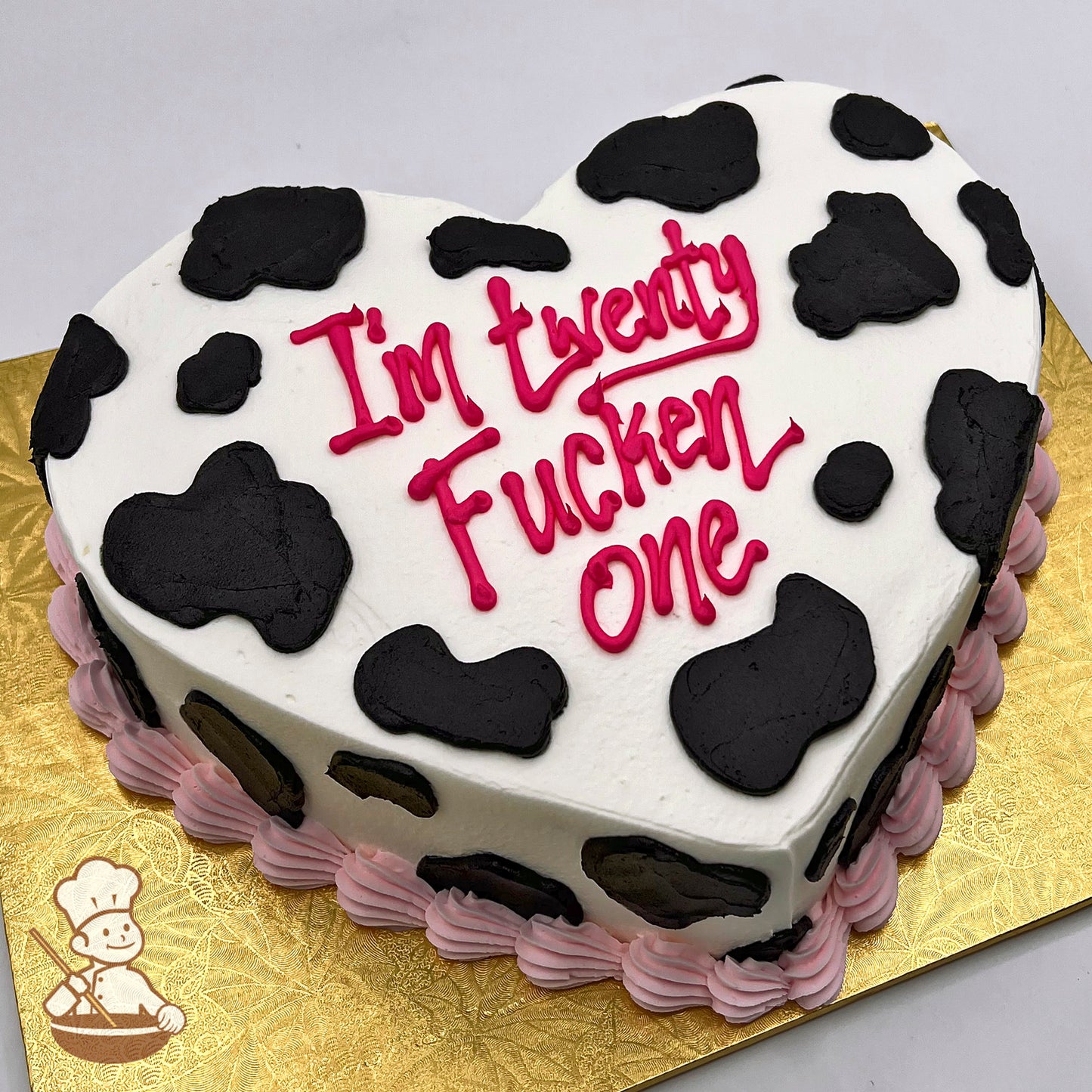Heart-shaped cake with black cow print buttercream spots, white frosting base, and pink piped border on a gold board.