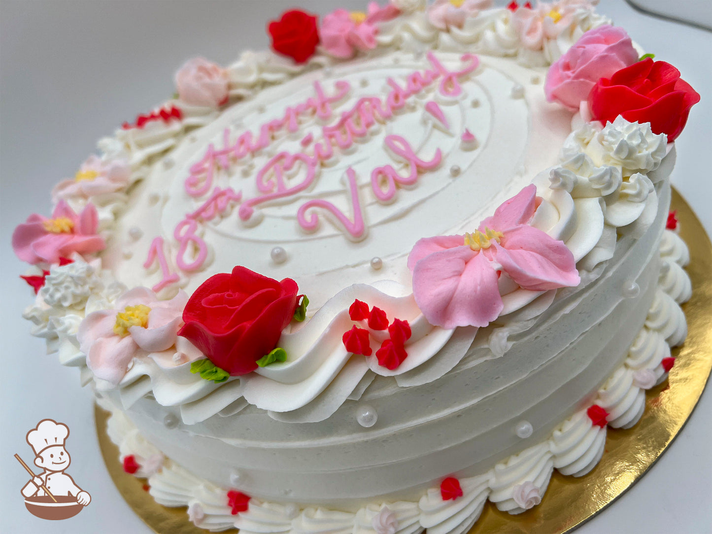 Round cake with white frosting, decorated with pink and red buttercream flowers, small pearls, and piped swirl trims.