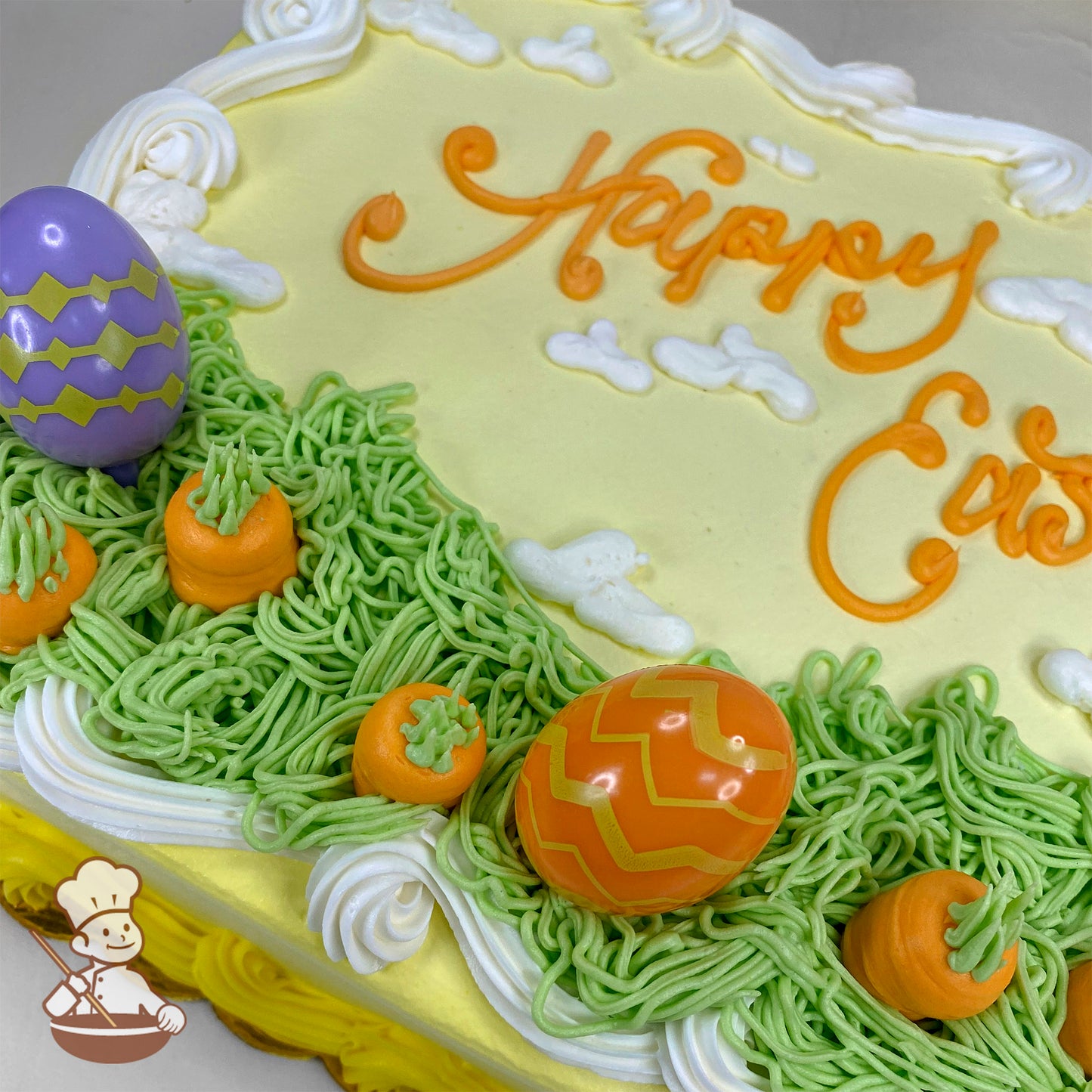 Sheet cake with yellow icing, white clouds, green piped grass, orange buttercream carrots, and plastic Easter egg toppers in orange and purple.