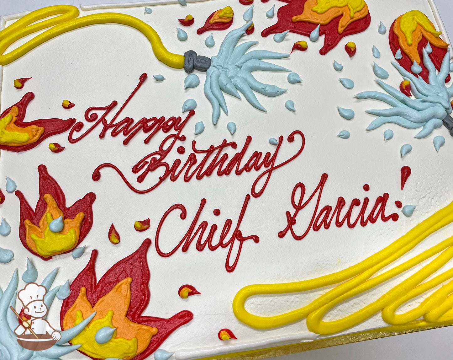 Sheet cake with buttercream decorations, featuring red and yellow flames, blue water splashes, a yellow hand-piped hose, and scattered water droplets.