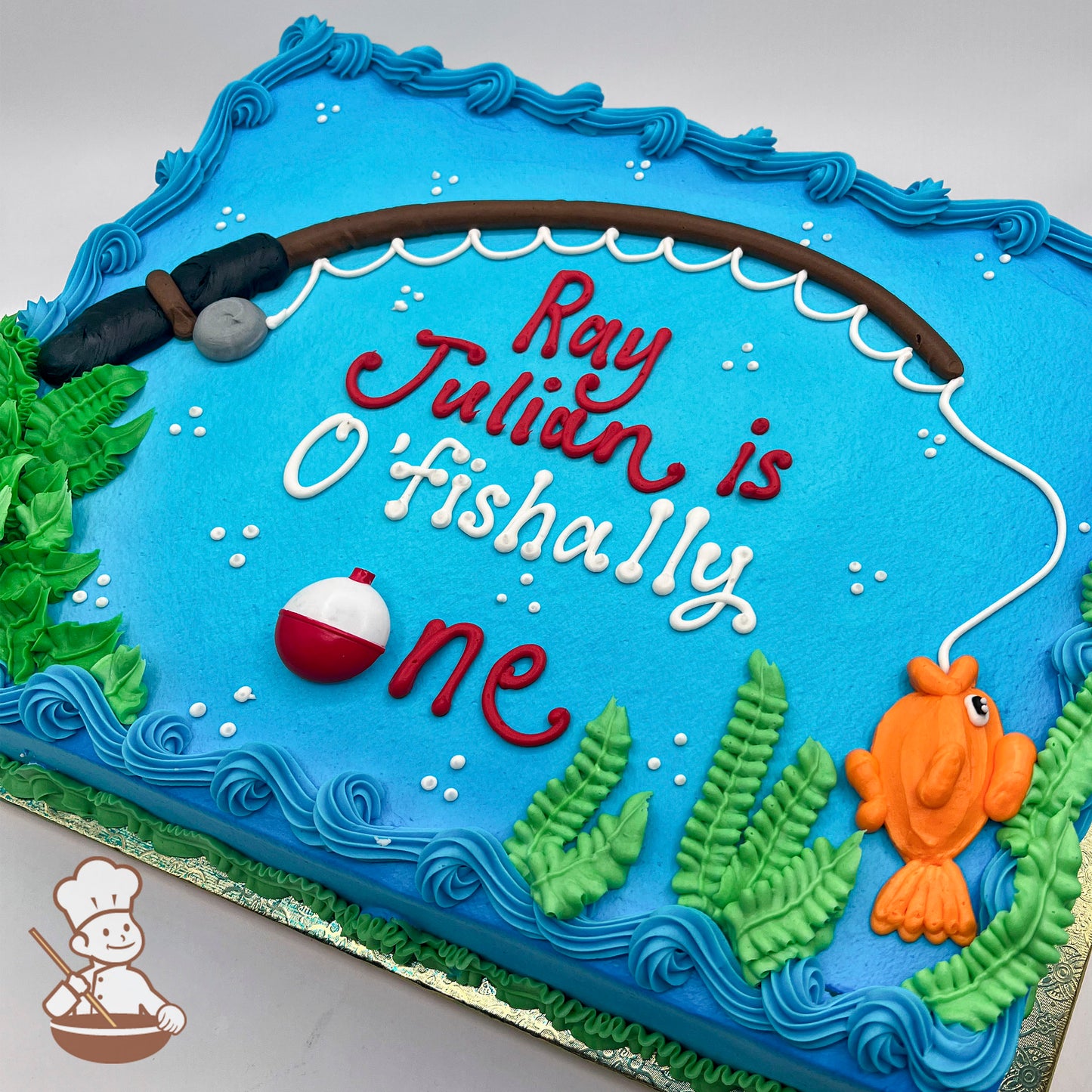 Sheet cake with blue frosting, a hand-piped fishing rod, a bobber, green seaweed, and an orange fish on the line.