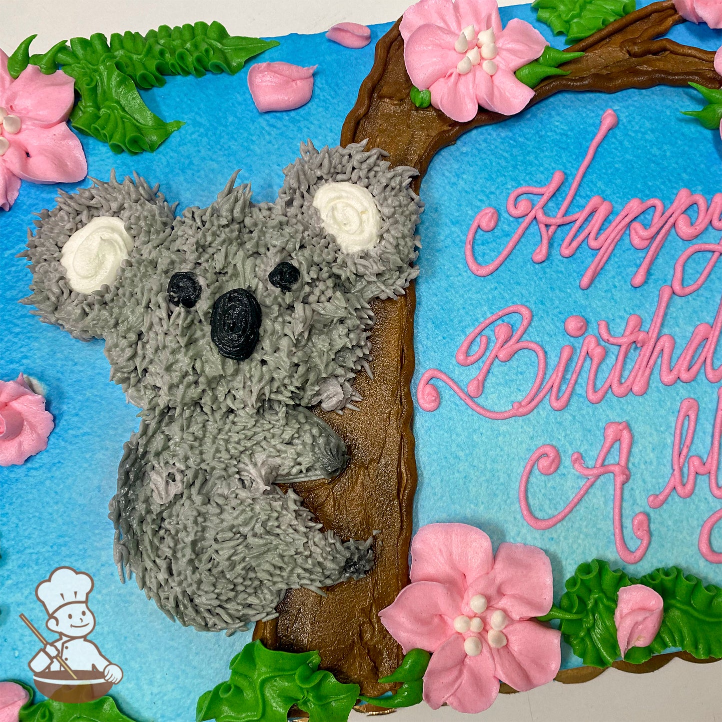 Gray hand-piped koala on a brown branch, surrounded by pink buttercream blossoms and green leaves on a blue background.