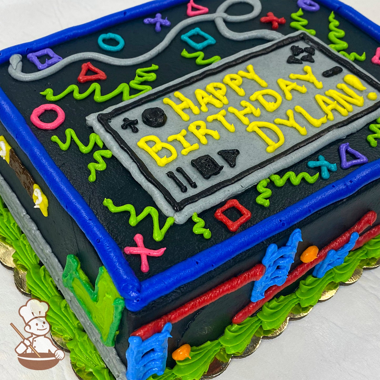 Sheet cake with black icing base, decorated with colorful PlayStation controller symbols (circle, square, triangle, X), green squiggles, and a gray controller design on top.