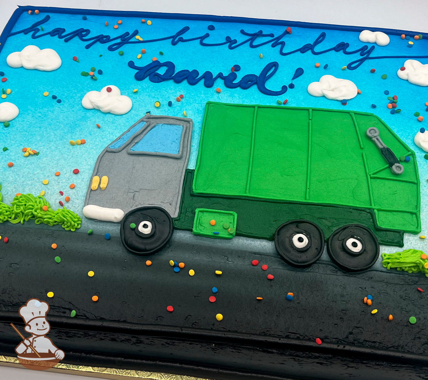 Sheet cake with icing decorations of a green garbage truck on a black road, blue sky, white clouds, and colorful sprinkles scattered throughout.