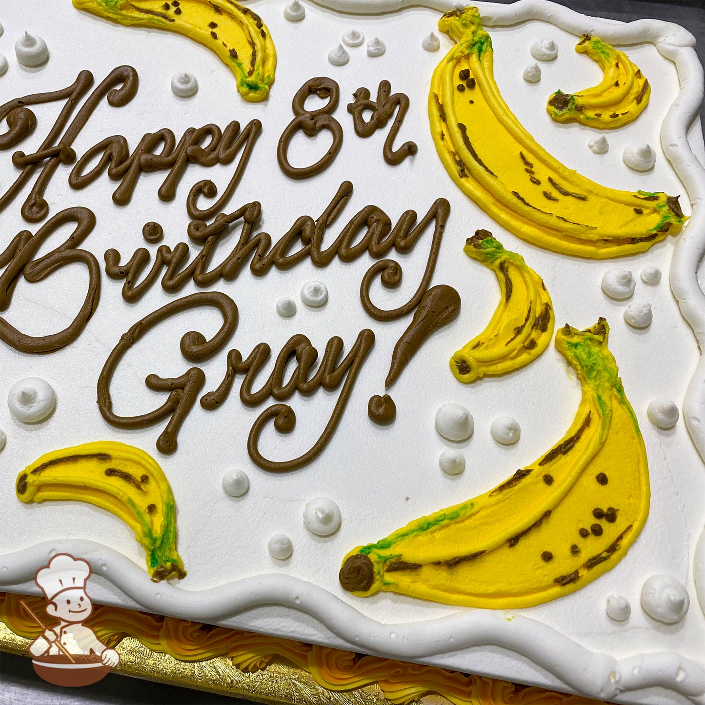 Sheet cake with white frosting, decorated with yellow buttercream bananas, brown detailing, and small white buttercream dots.