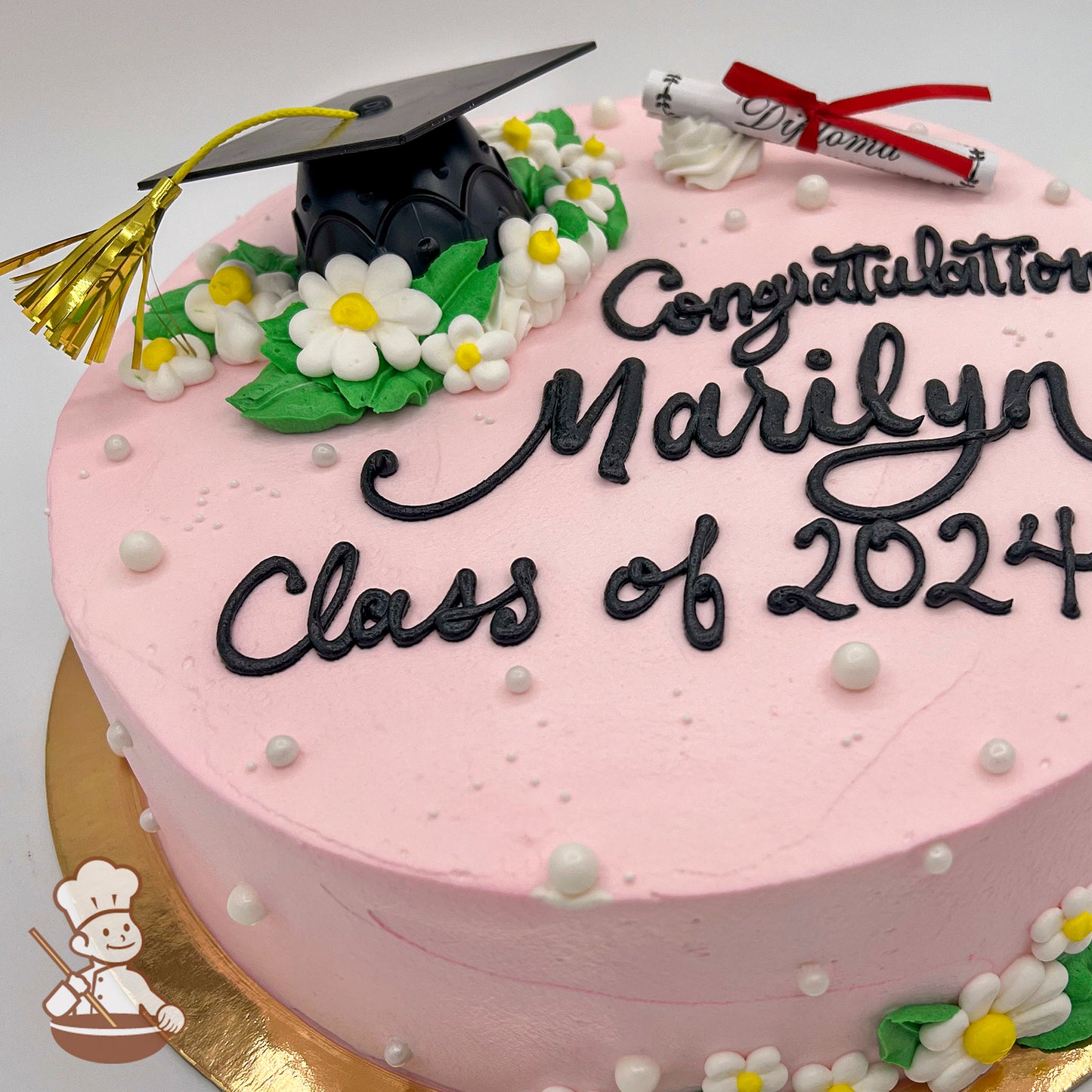 Round pink iced cake with white and yellow daisy decorations, green leaves, a plastic black graduation cap with a gold tassel, and a rolled paper diploma.