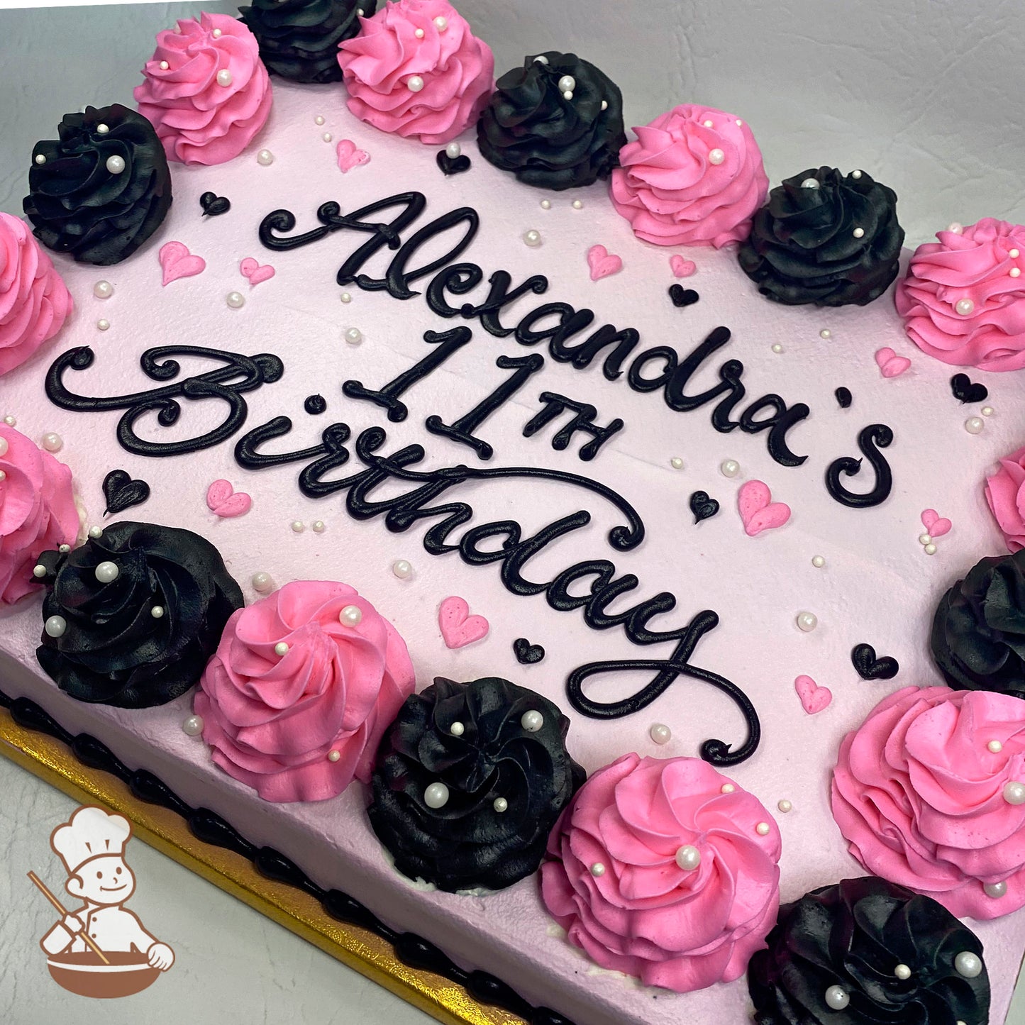 Pink and black buttercream swirls with sugar pearls and heart decorations on a sheet cake.