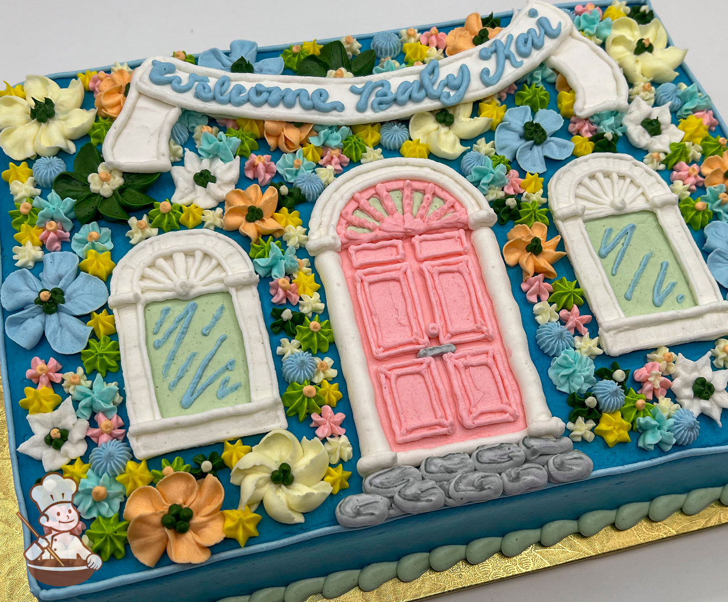 Sheet cake with buttercream decorations, featuring a pink door, two green-paned windows, colorful flowers, and a white banner on a blue background.