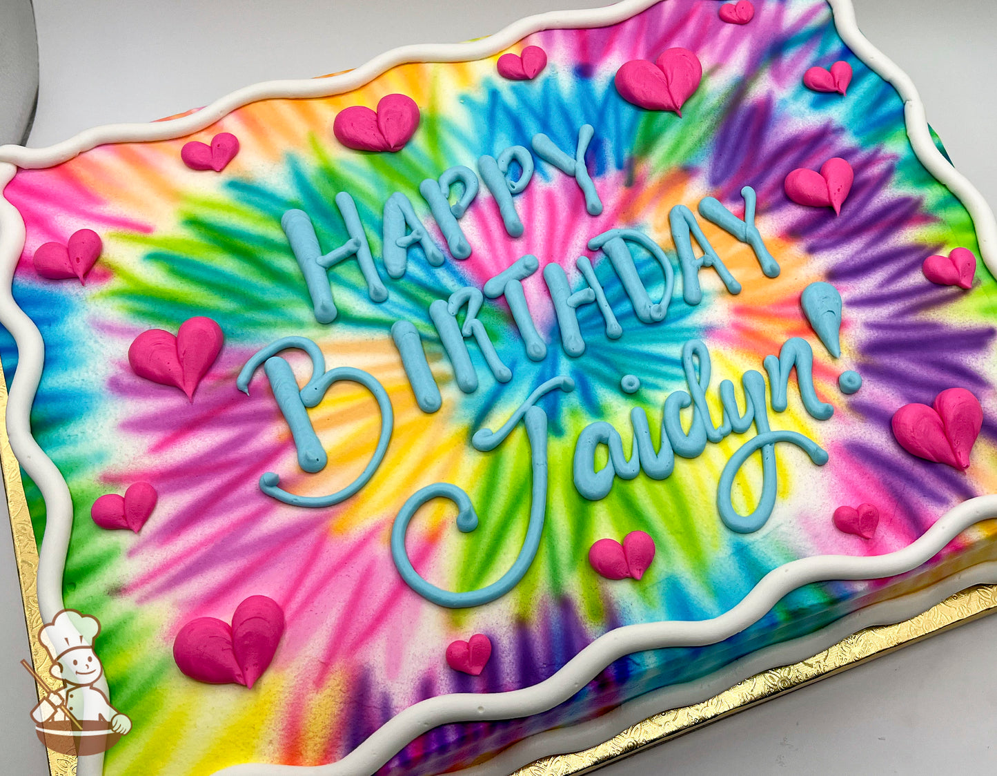 Colorful tie-dye sheet cake with vibrant swirls of pink, blue, yellow, green, and purple, decorated with pink buttercream hearts and a white border.