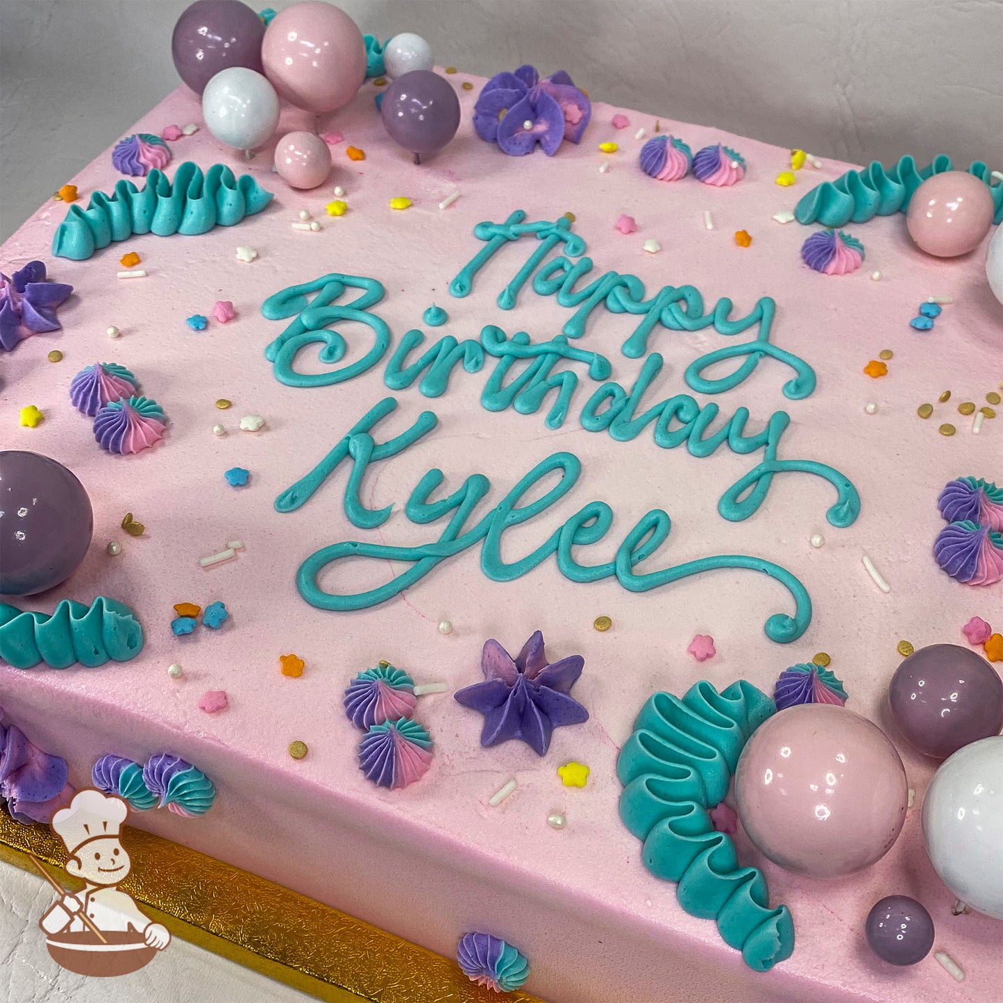 A pink sheet cake with teal buttercream swirls, purple and pink floral designs, colorful sprinkles, and plastic bubble toppers in pink shades.