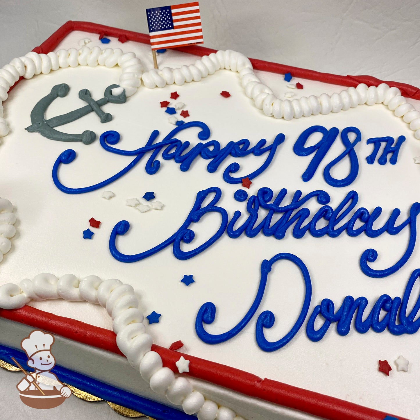 Sheet cake with white frosting, a gray anchor design, a small American flag, red and blue borders, and star-shaped patriotic sprinkles.