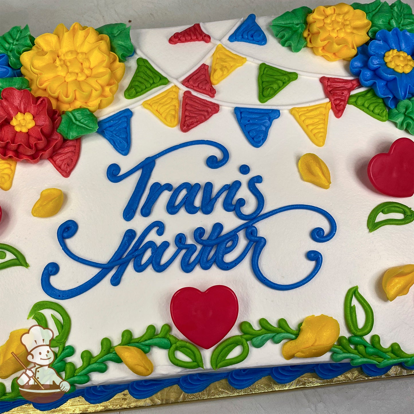 Sheet cake with white frosting, colorful buttercream party banners, yellow and red camellia flowers, green leaves, and red plastic heart decorations.