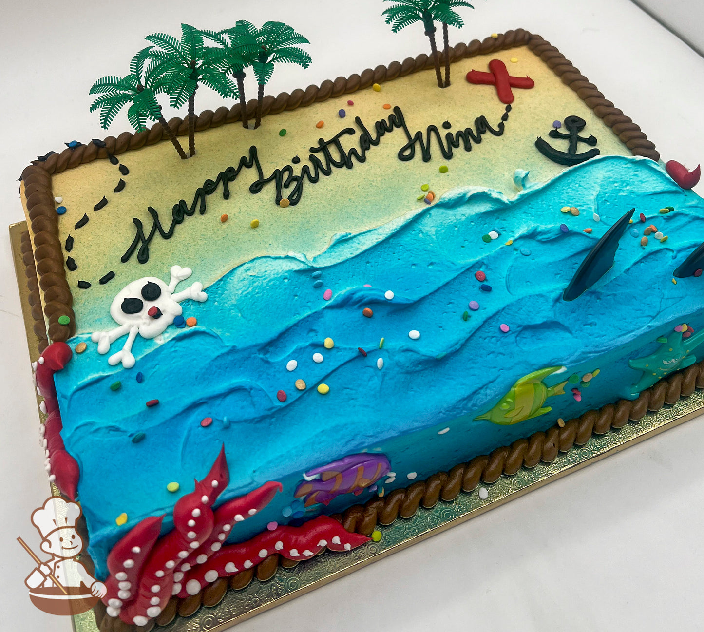 Sheet cake with buttercream decorations featuring a beach and ocean theme, with plastic palm trees, a red "X," an anchor, a skeleton head, plastic shark fins, fish, and a starfish. Rope trim borders the cake.
