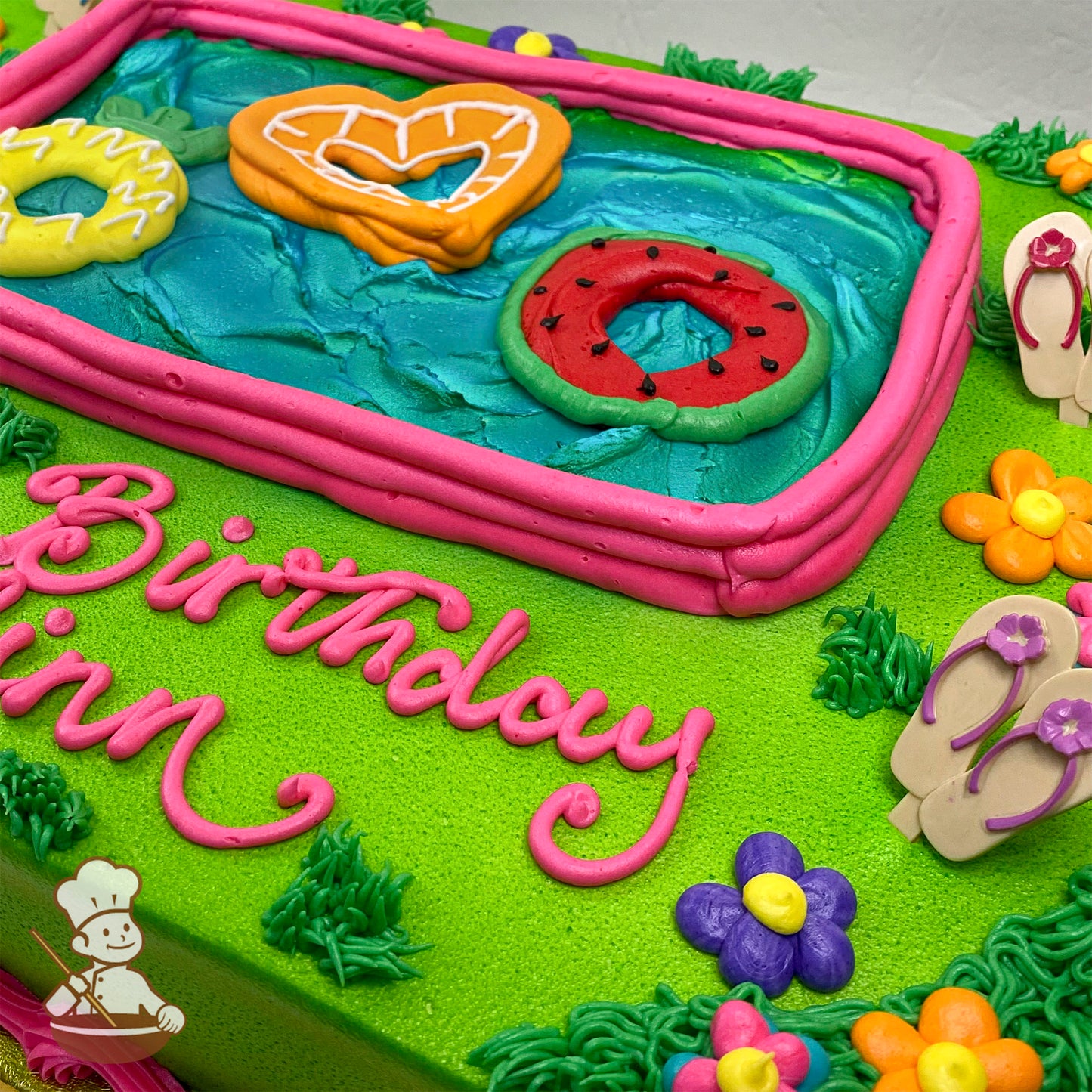 Sheet cake with a buttercream pool, pink border, blue water, floaties shaped like a heart, pineapple, and watermelon, surrounded by grass, flowers, and plastic sandal picks.