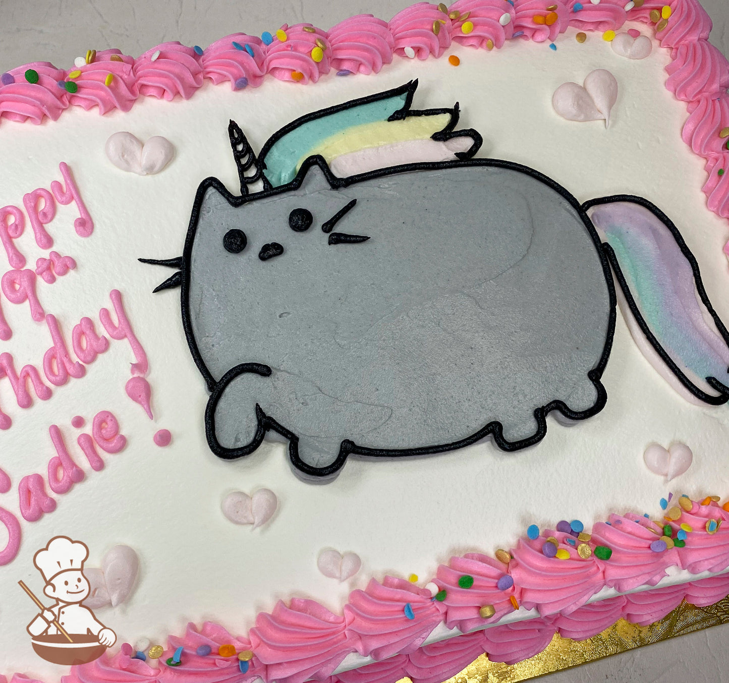 Sheet cake with a buttercream Pusheen cat design as a unicorn, pastel rainbow mane and tail, pink frosting border with sprinkles, and small pink hearts.