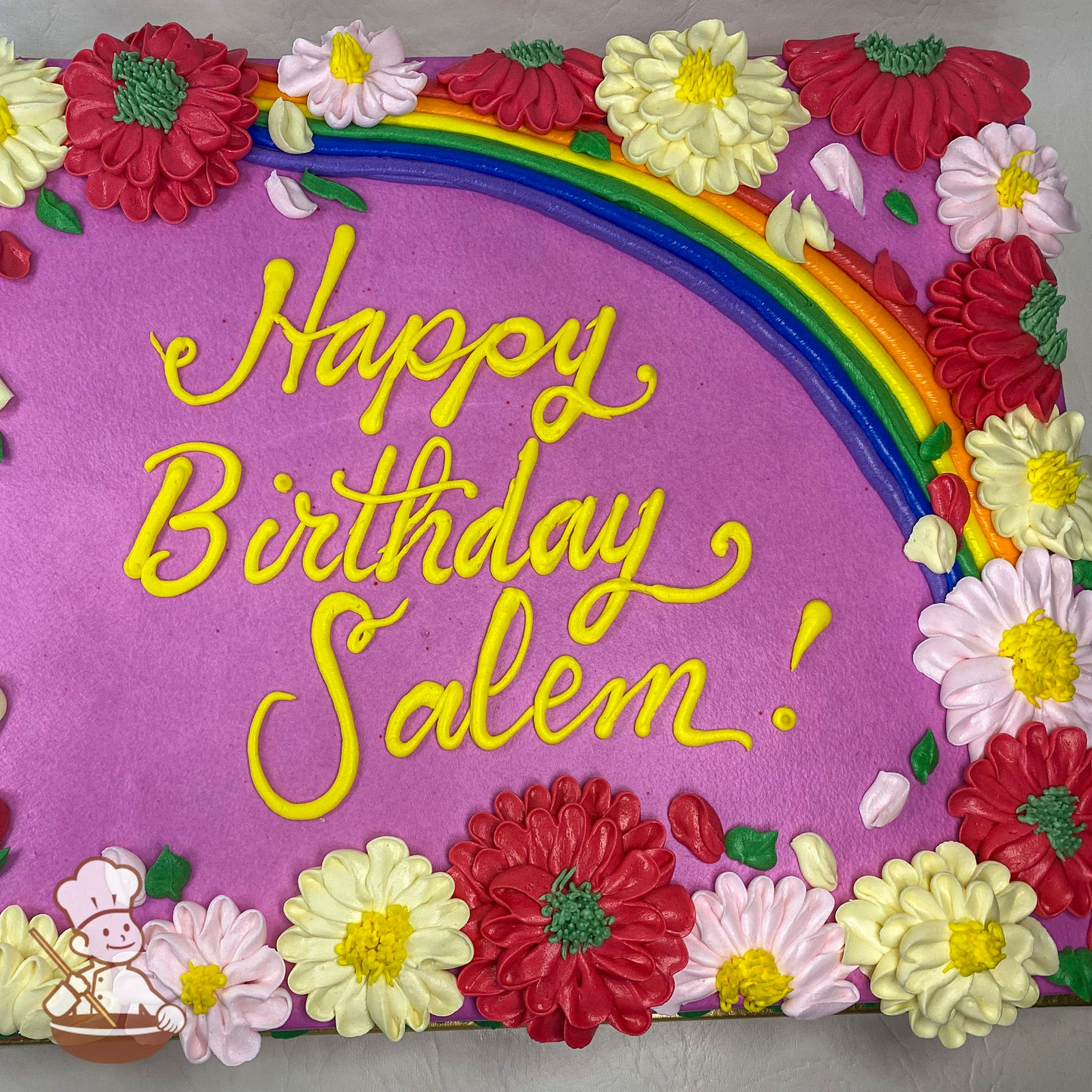 Purple sheet cake with a rainbow decoration and buttercream flowers, including red, yellow, and pink mums, arranged around the edges.