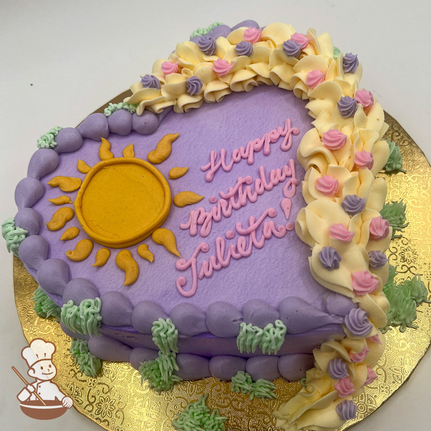 Heart-shaped sheet cake with purple buttercream, a yellow sun design, a braided buttercream border with pink and purple rosettes, and green grass accents.