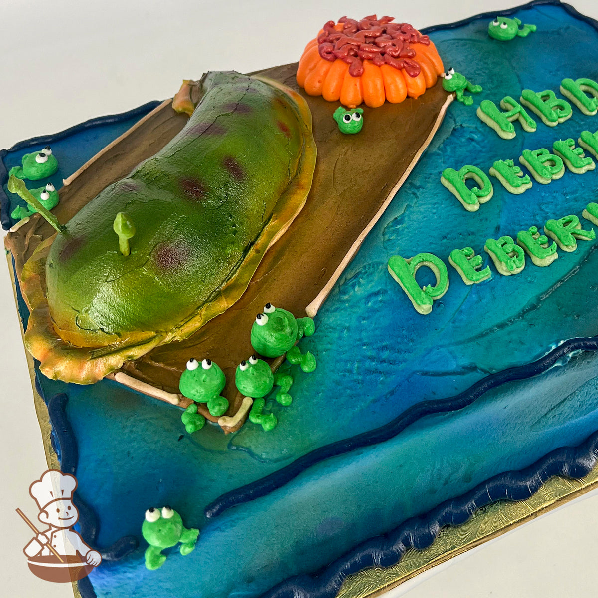 A sheet cake with a green slug-shaped buttercream decoration on a brown platform, surrounded by small green frog figures and blue water-like icing.