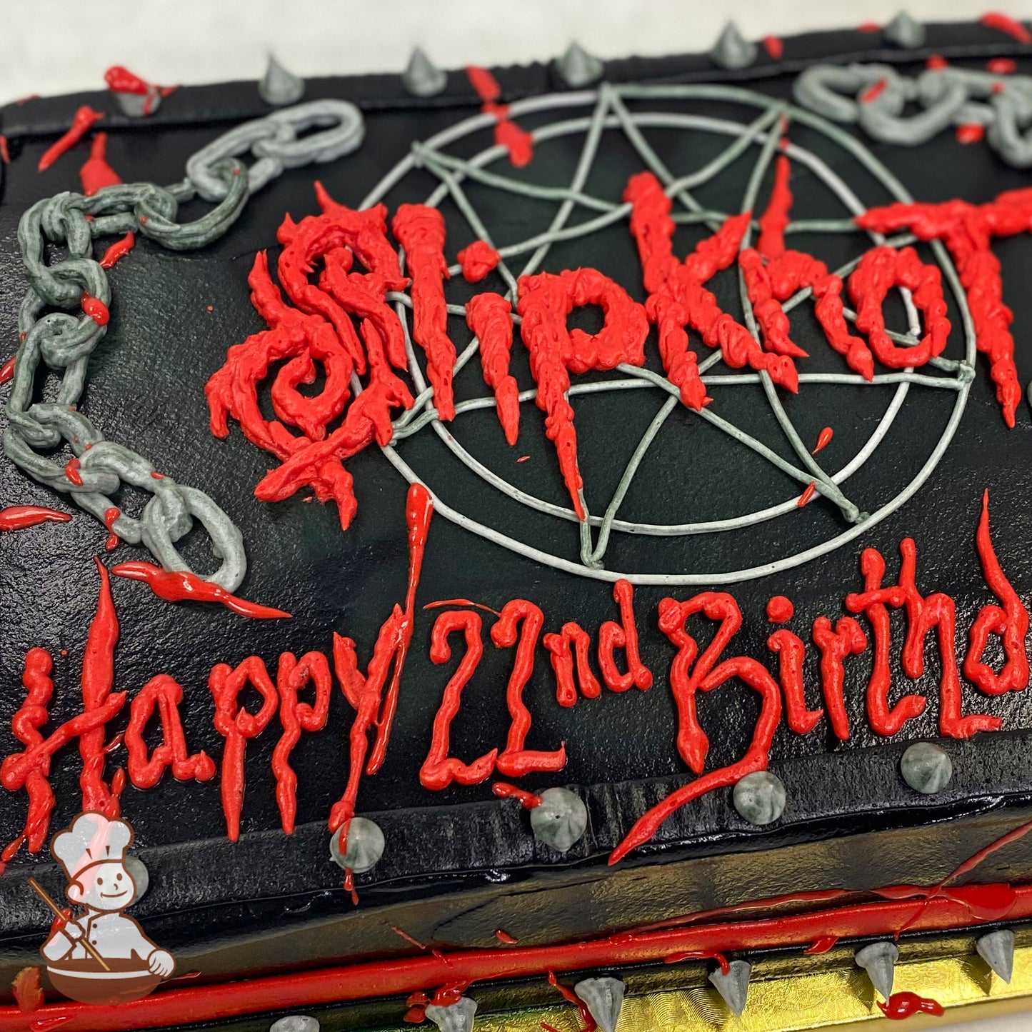 A black sheet cake with red buttercream text, a pentagram design, gray buttercream chains, and spikes along the edges for decoration.