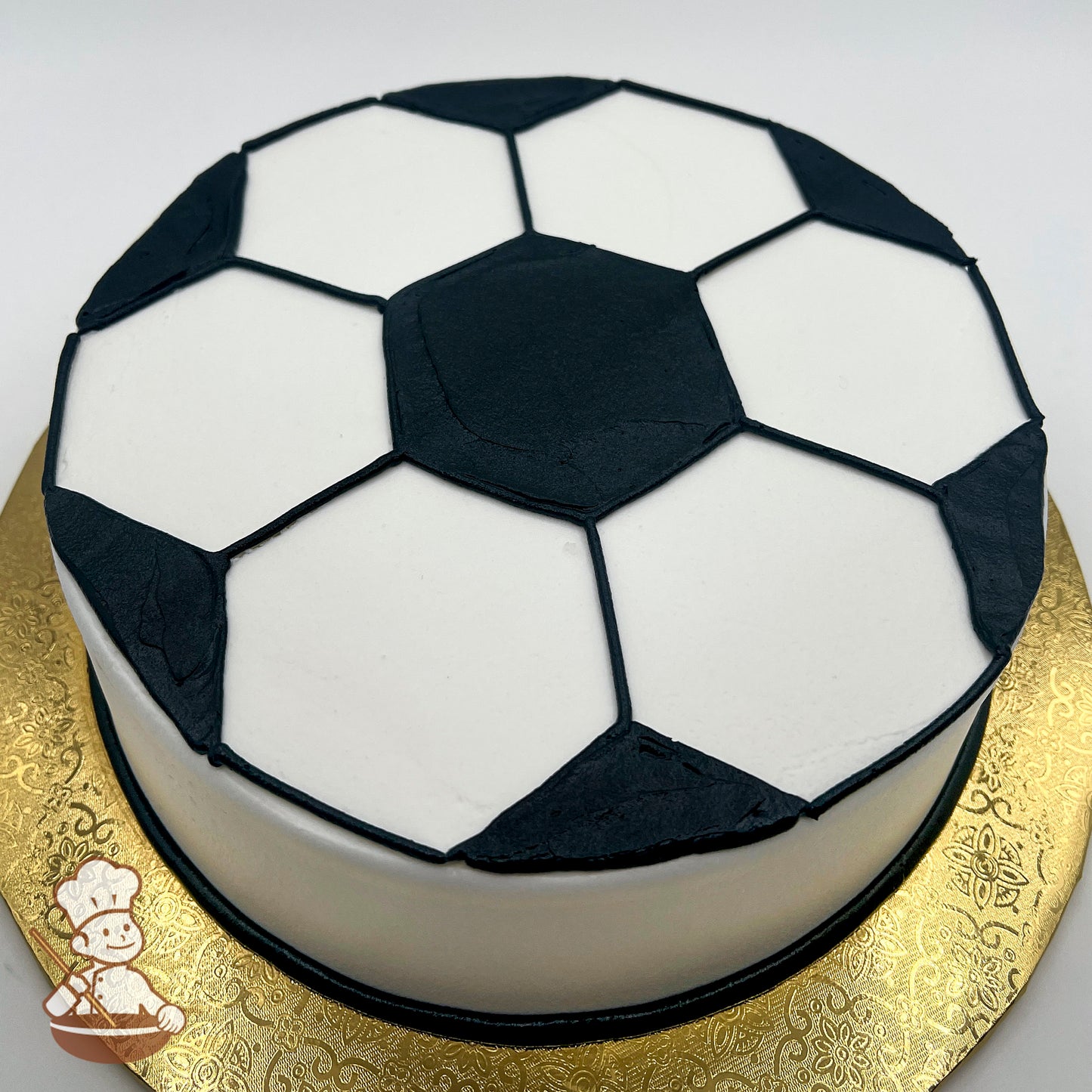 Round cake decorated like a soccer ball with black and white buttercream hexagons, on a gold patterned board.