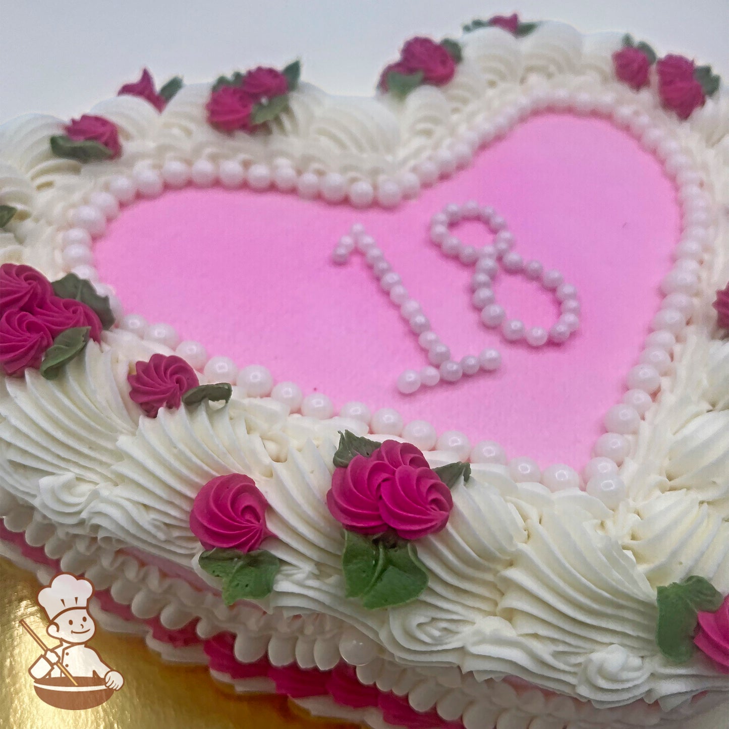 Heart-shaped cake with white buttercream swirls, pink flowers, green leaves, and sugar pearl number "18" on a pink background.