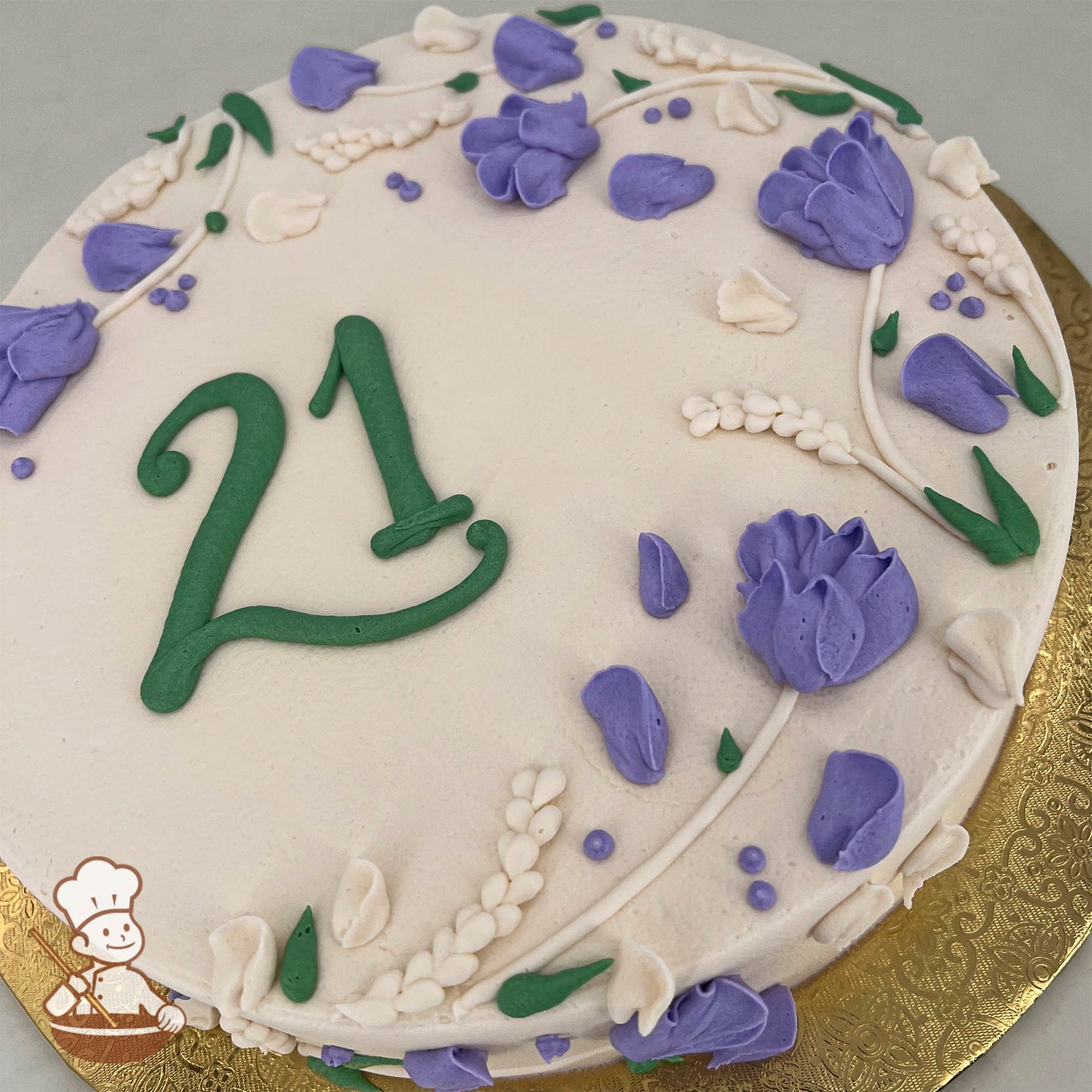 Round sheet cake with white frosting, decorated with purple buttercream flowers, green leaves, and white floral accents on a gold base.