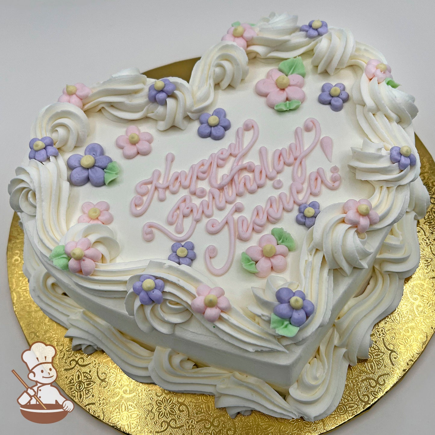 Heart-shaped cake with white buttercream swirls, pastel pink and purple daisy decorations, and green leaves on a gold board.