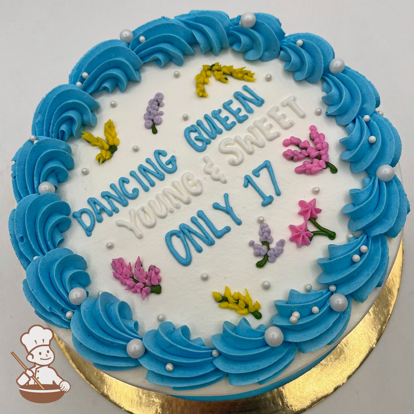 Round cake with blue buttercream swirls on the border, decorated with small sugar pearls and buttercream flowers, including pink, yellow, and purple blossoms.