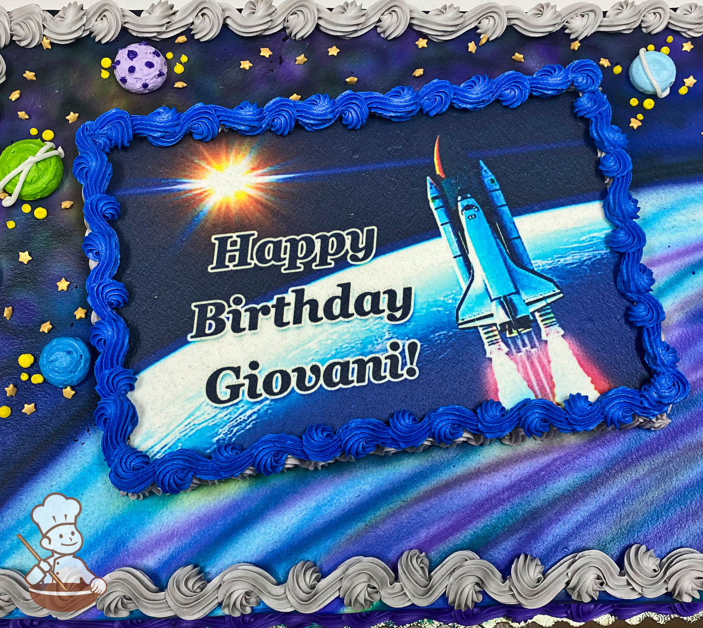 Sheet cake with a galaxy-themed design, featuring blue and gray piped borders, buttercream planets, and star-shaped sprinkles around the edges.