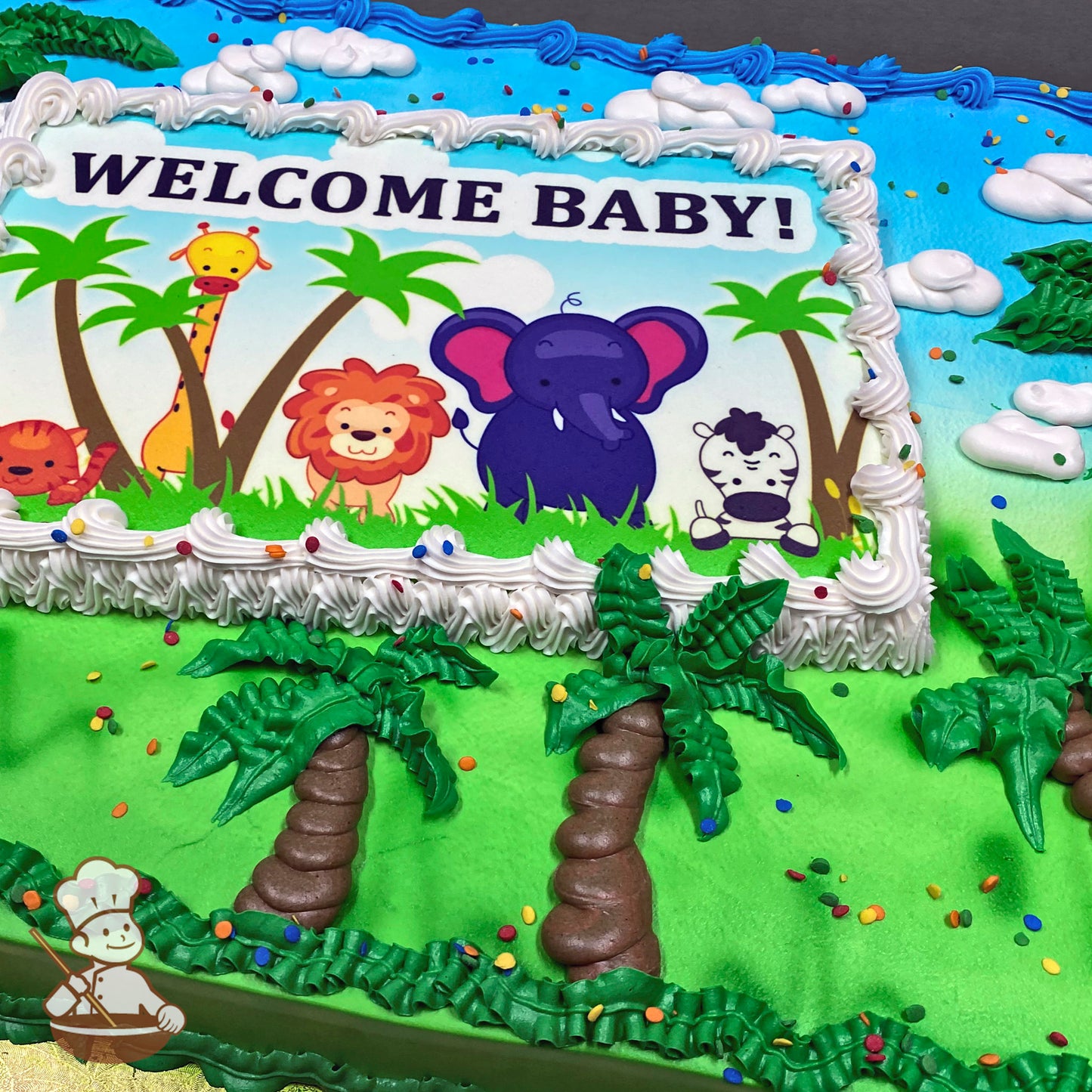 Sheet cake with green grass, piped palm trees with brown trunks and green leaves, colorful sprinkles, and a white border around a printed image.