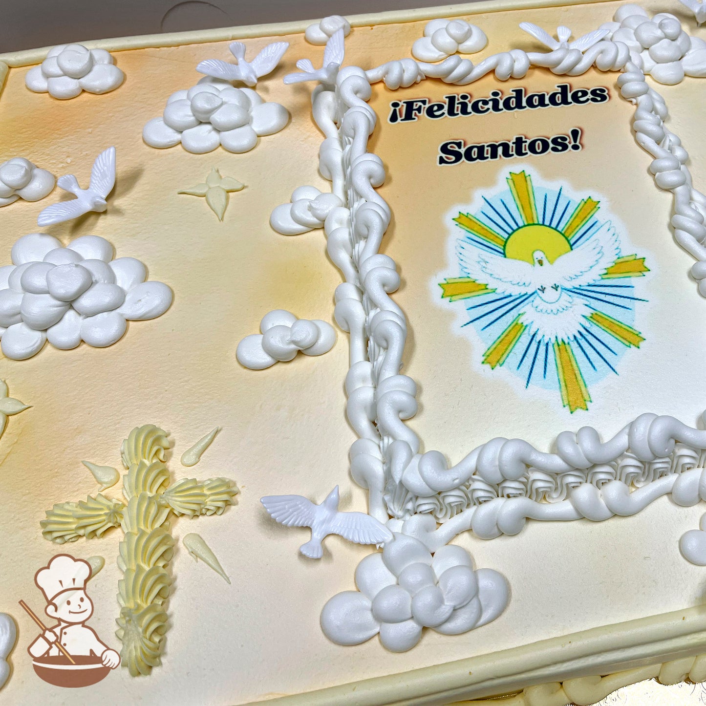 Sheet cake with buttercream decorations, featuring a scallop cross, white piped clouds, and plastic doves. A printed image is framed with piped borders.
