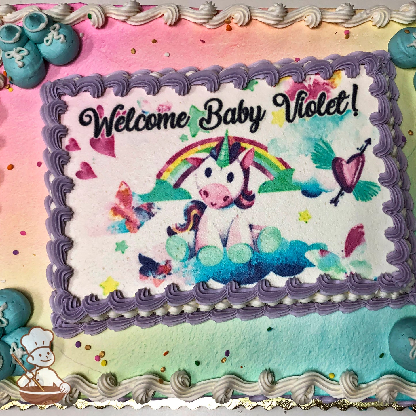 Sheet cake with pastel rainbow spray background, purple piped border, aqua buttercream baby booties, and colorful confetti accents.