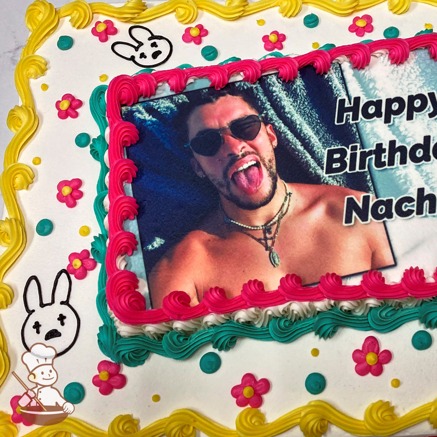 A sheet cake with colorful buttercream decorations, including yellow, pink, and teal borders, small pink and yellow flowers, and black bunny face designs.