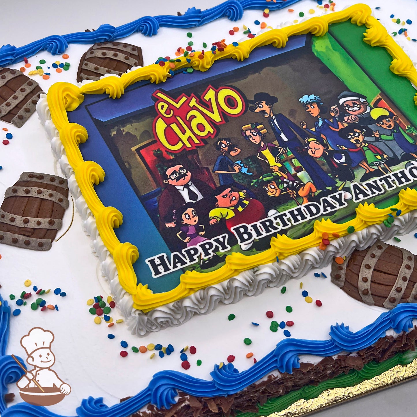 Sheet cake with a printed image surrounded by yellow buttercream borders, blue and white piped edges, colorful sprinkles, and barrel-shaped decorations.