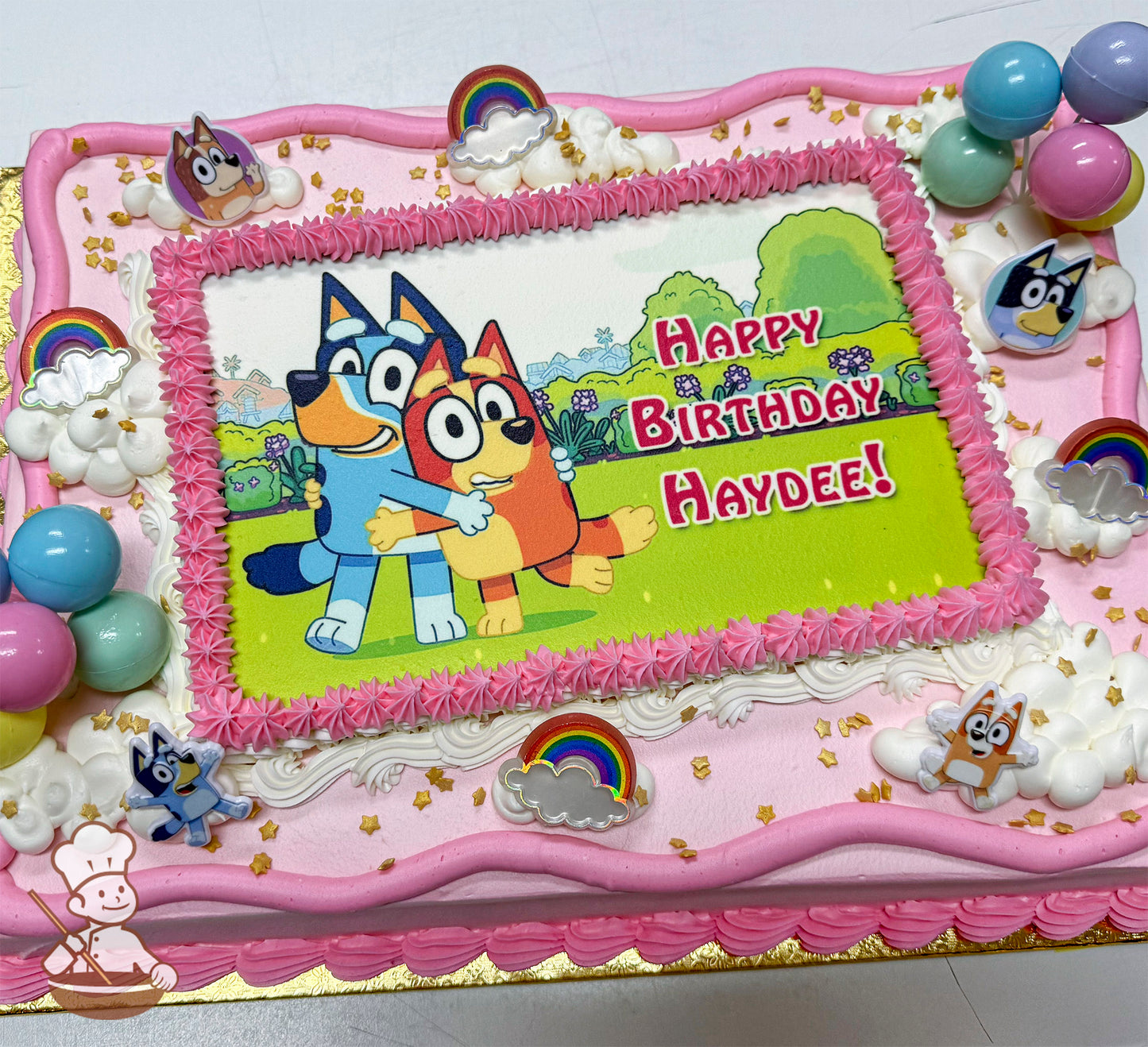 A rectangular sheet cake with pink and white buttercream borders, decorated with plastic Bluey character toys, rainbow toppers, hand-piped clouds, and gold star sprinkles.