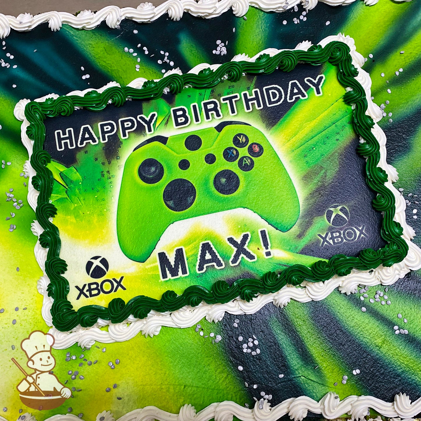 Sheet cake with green and black spray design, bordered by white and green buttercream swirl trims, silver confetti sprinkles scattered on top.