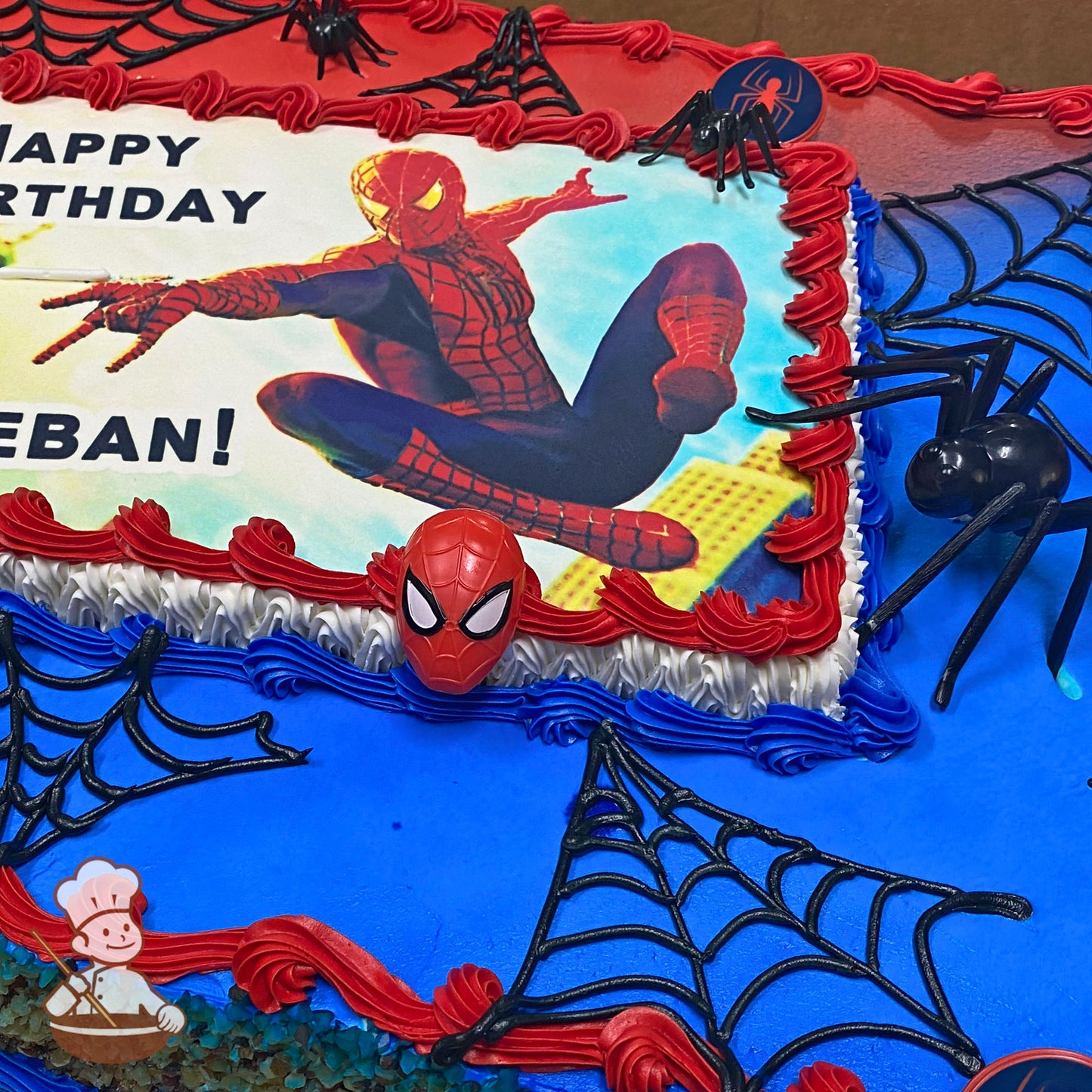 Sheet cake with blue and red buttercream borders, black spider web designs, plastic spiders, and a Spider-Man mask decoration.