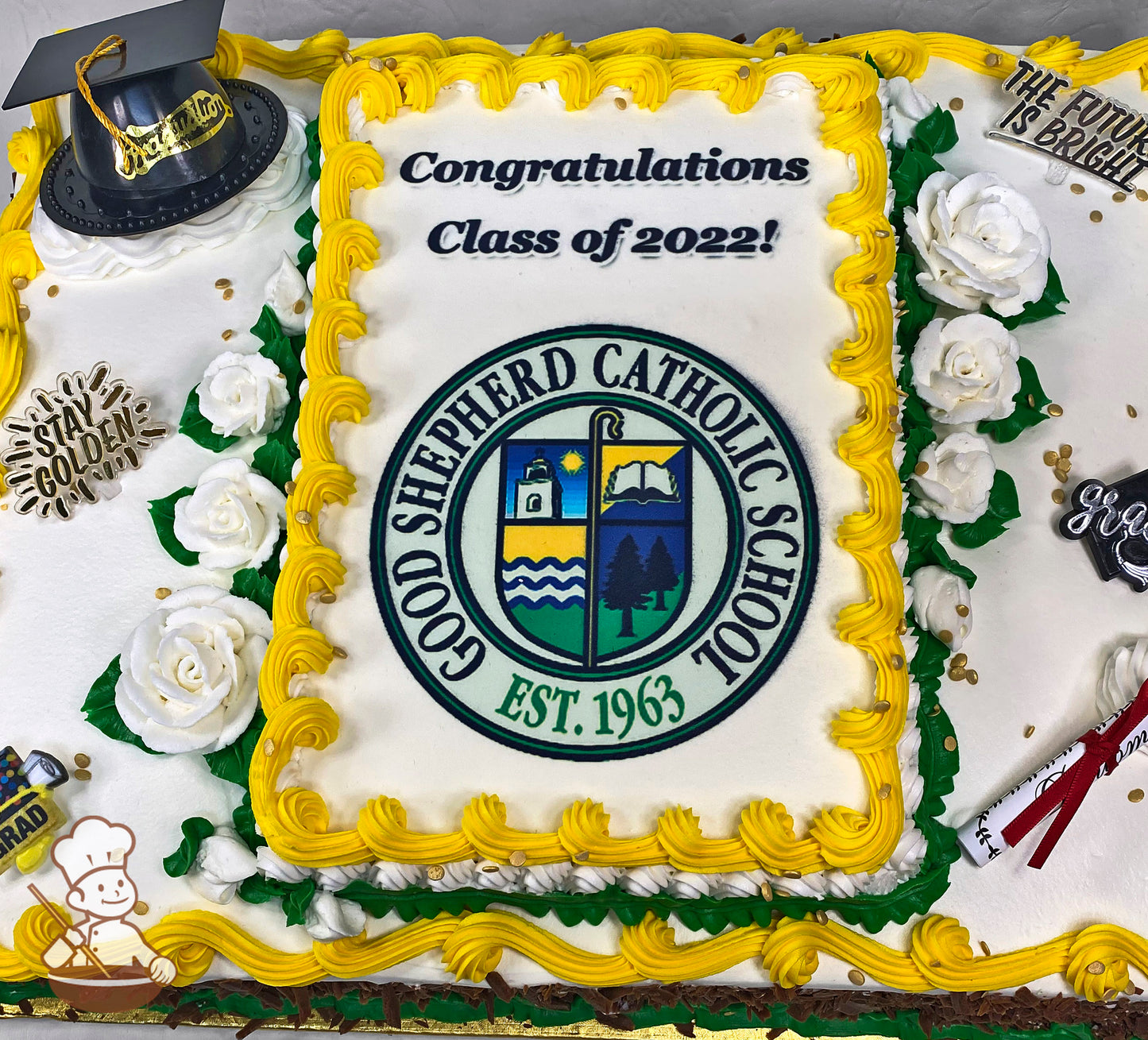 Sheet cake with yellow buttercream borders, white buttercream roses with green leaves, and gold accents. Graduation-themed decorations are placed around the cake.
