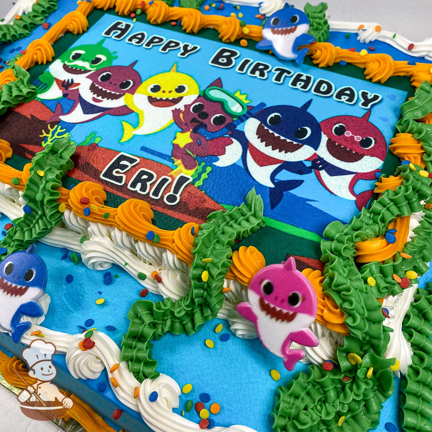 Sheet cake with a printed photo layer, surrounded by buttercream kelp, orange and white swirl trims, and plastic Baby Shark toys.