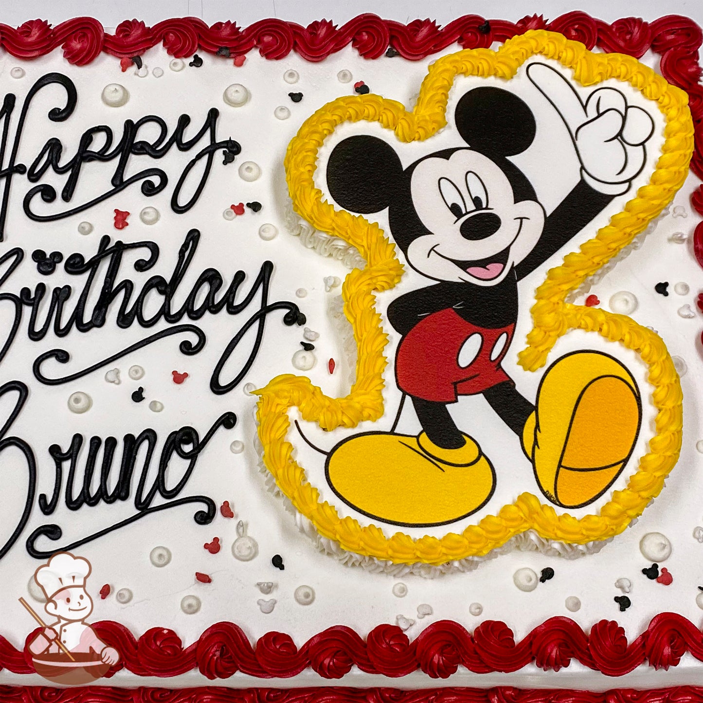 Sheet cake with white frosting, red border, and a Mickey Mouse design outlined in yellow frosting. Scattered small Mickey-shaped sprinkles.