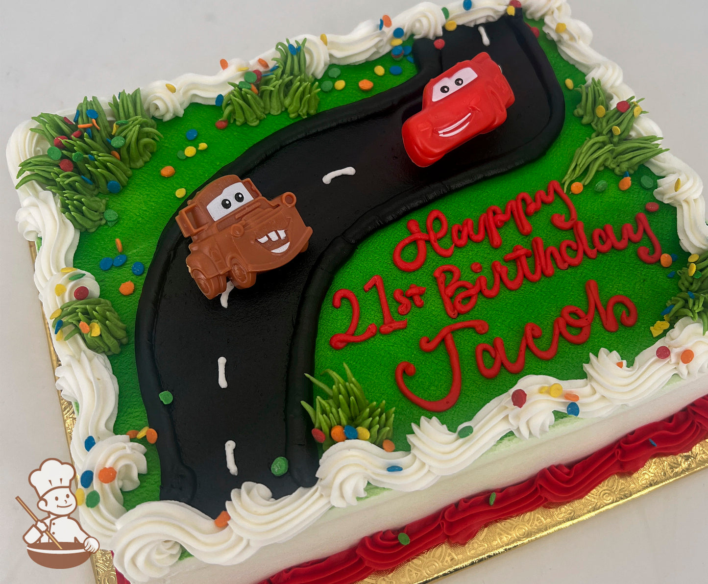 Sheet cake with a green base, a black road design, plastic Lightning McQueen and Mater toys, white frosting border, and colorful sprinkles.