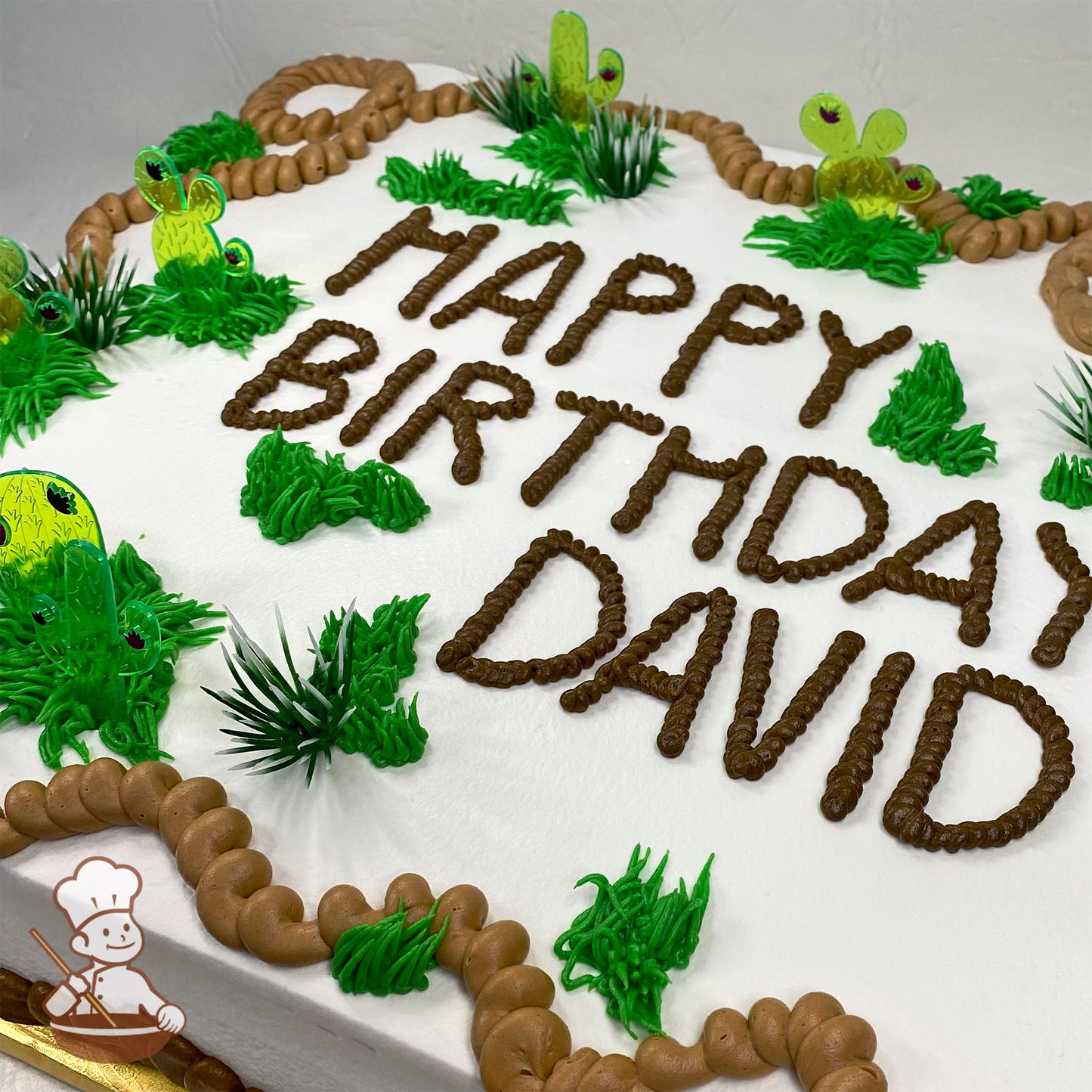 Sheet cake with white frosting, decorated with plastic cactus toppers, green buttercream bushes, and brown buttercream rope trim.