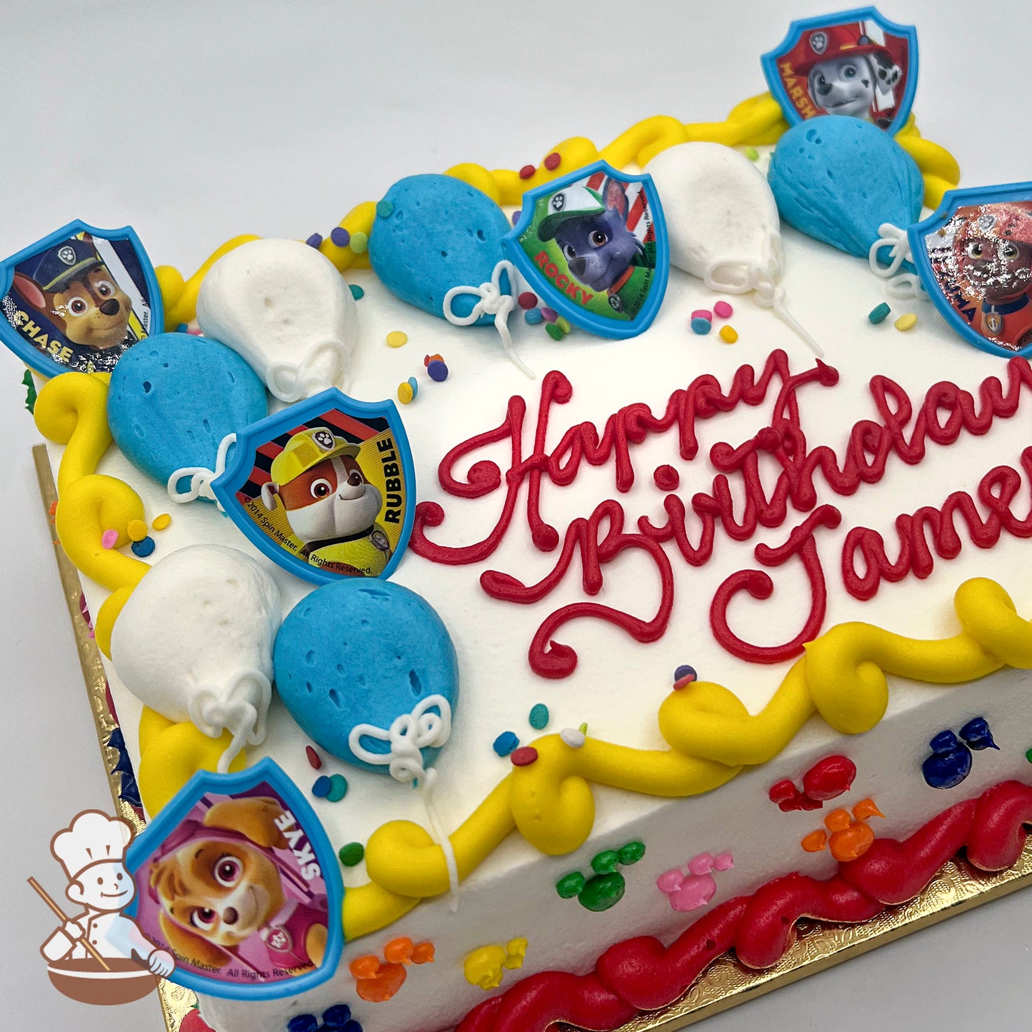 Sheet cake with white frosting, yellow and red piped borders, colorful paw prints, blue and white balloon decorations, and Paw Patrol character plastic rings.