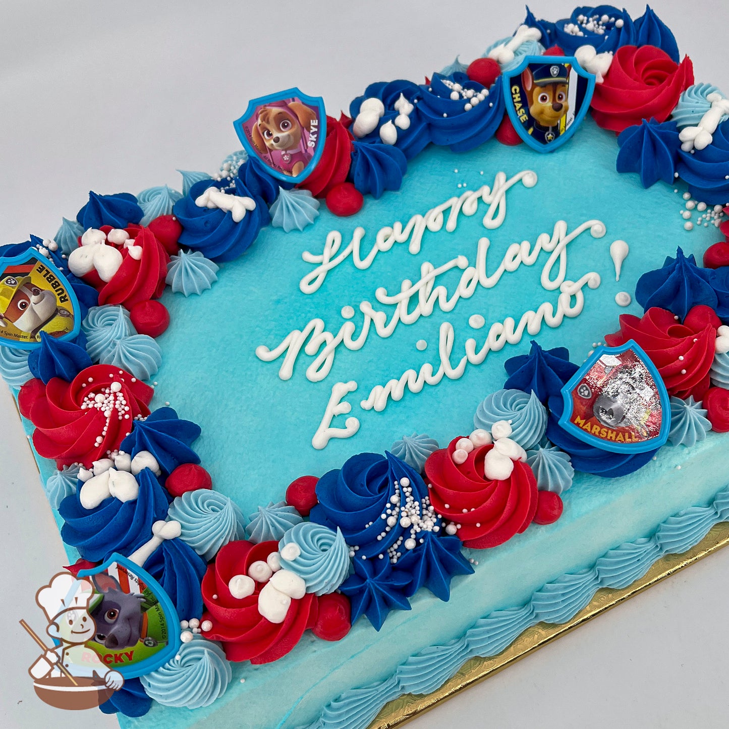 Sheet cake with light blue frosting, red and blue swirled borders, white pearl sprinkles, and Paw Patrol plastic rings featuring characters like Skye and Chase.
